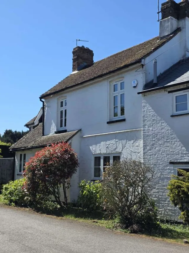 A weathered tiled roof crowns a white cottage behind flowering shrubs and a low wall.