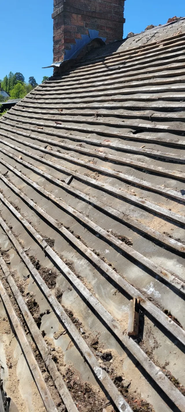 Uneven old battens cross a stripped roof slope below a brick chimney.