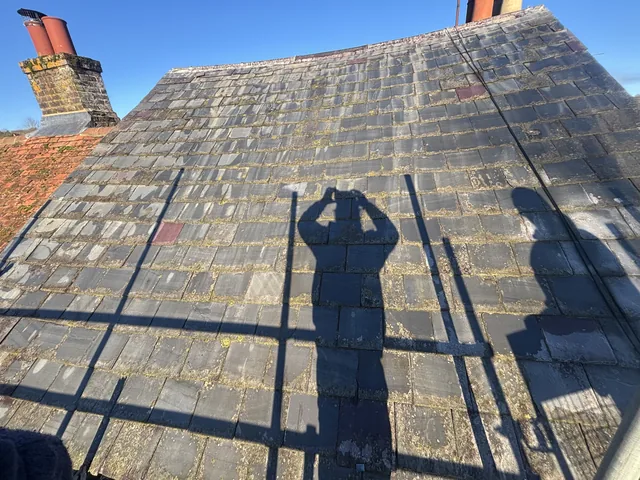 Weathered slate courses carry scaffold shadows between brick chimneys beneath a clear blue sky.