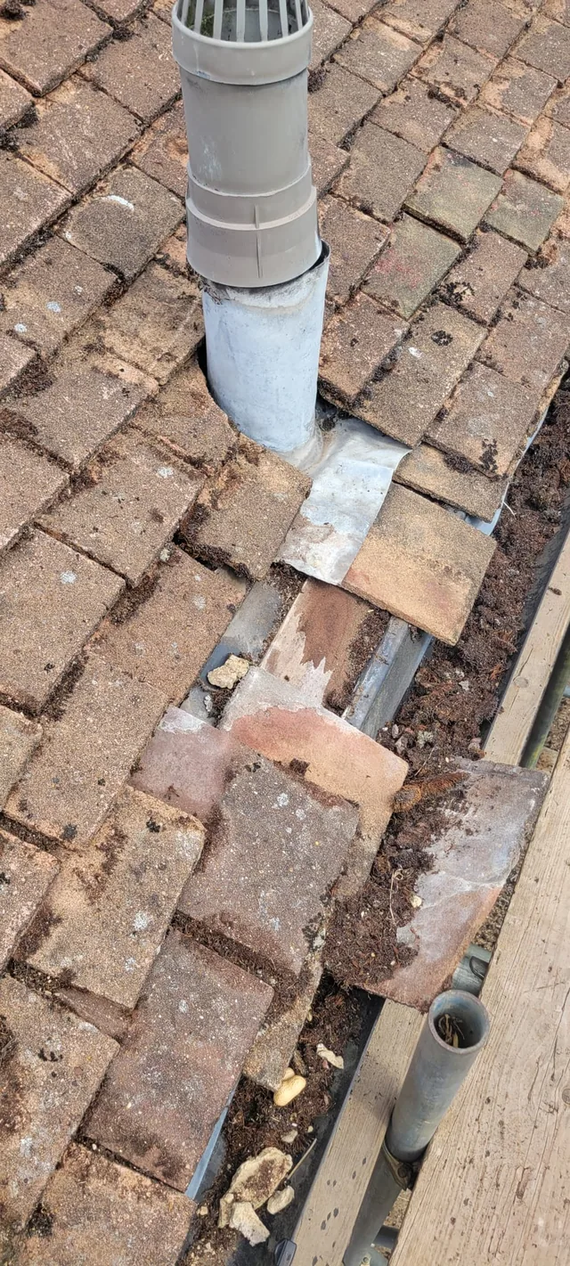 A capped flue stands above old flashing beside missing tiles and debris along a hip.