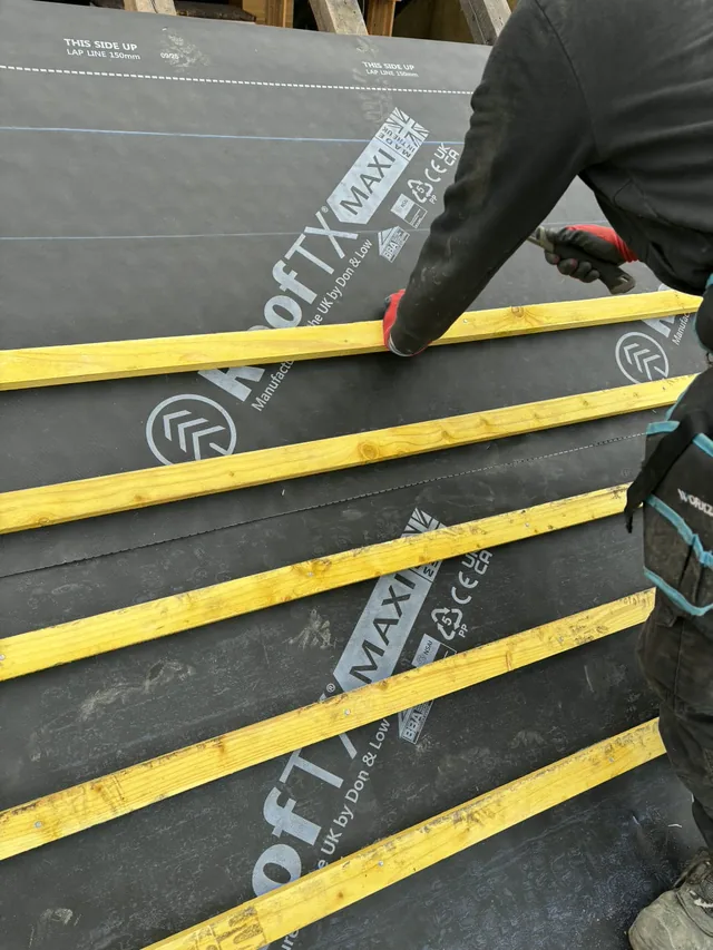 A roofer fixes a yellow batten across dark membrane using a hand tool.