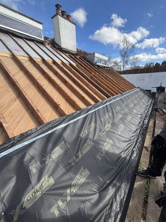A broad roll of dark membrane covers lower rafters beneath rooflights and a chimney.