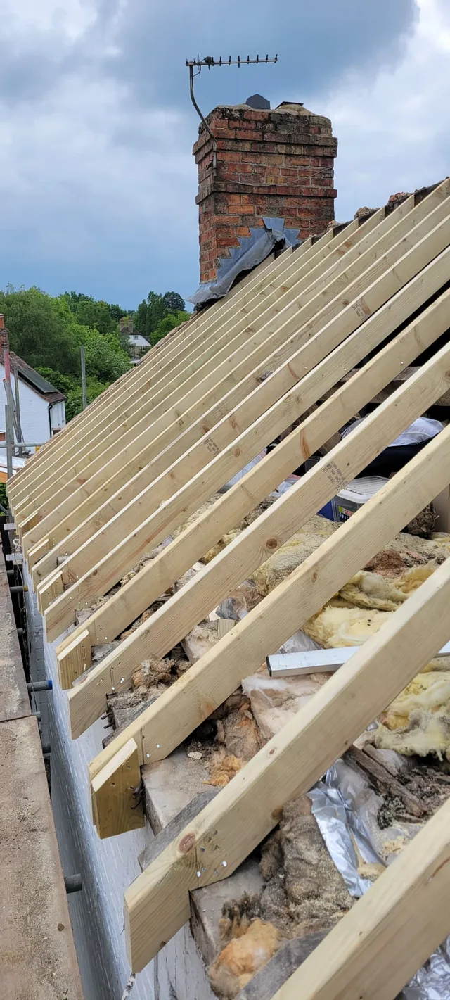 New pale rafters rise above loose insulation towards a brick chimney and aerial.
