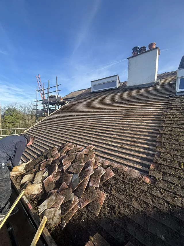 A roofer stacks removed tiles below exposed battens and a white chimney.