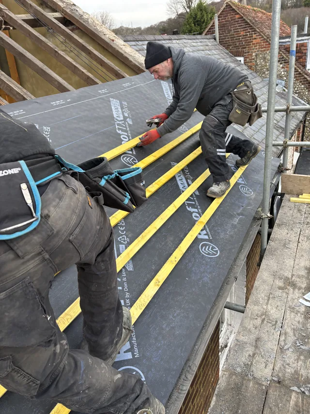 Two roofers fit yellow battens over dark membrane beside a scaffold platform.