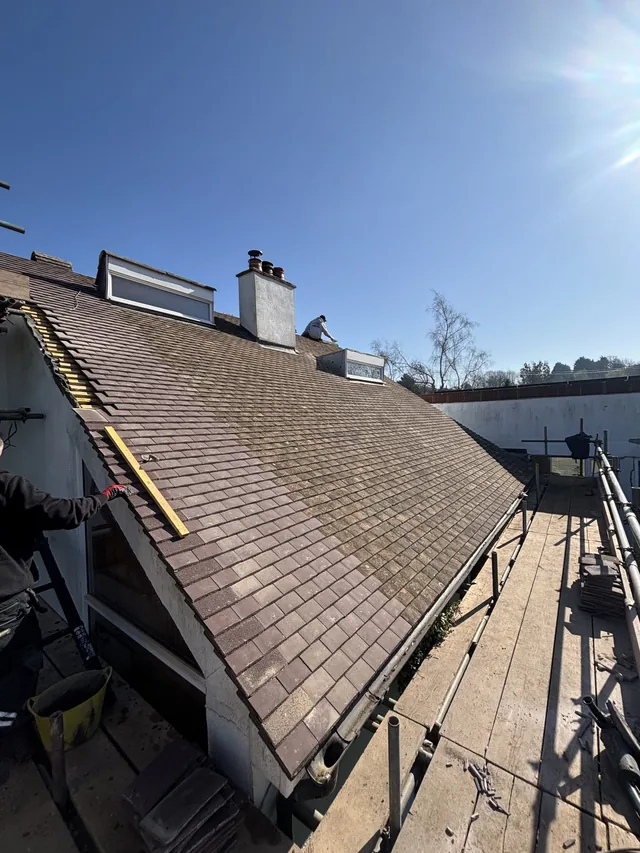 Roofers finish laying brown tiles around a white chimney and two raised rooflights.