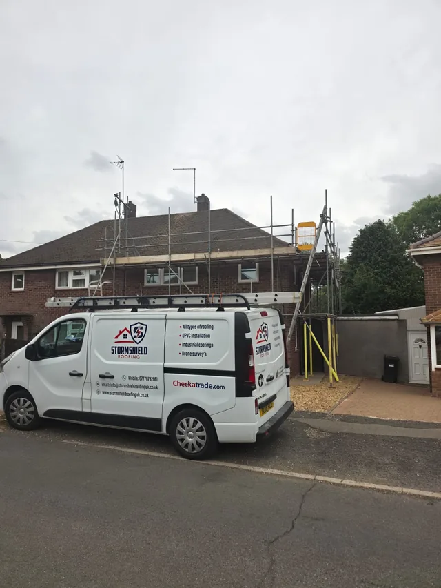 A white Stormshield van with roof ladders stands outside a scaffolded brick house.